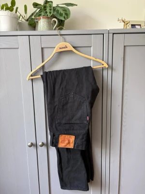 Svarta Levi's 501 jeans 30/34 - Svarta klassiska Levi's 501 jeans i rak modell med fem fickor och knappgylf. Jeansen har den ikoniska läderpatchen bak i midjan och röd Levi's-tag på bakfickan. Tillverkade i bomull och passar perfekt till en avslappnad streetstil.