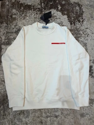 Vit sweatshirt från Prada - Säljer en stilren vit sweatshirt från Prada med rund halsringning och diskret röd logga på bröstet. Tröjan har en klassisk passform och är tillverkad i mjukt material som känns skönt mot huden. Perfekt för en clean och trendig look.