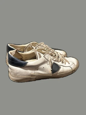 Vita och marinblå sneakers från Philippe Model - Snygga vita sneakers från Philippe Model med marinblå detaljer på häl och sidor. Skorna har klassisk låg profil, rund tå och vita snören. Tillverkade i läder med gummisula och ikonisk sköld på sidan. Perfekta för en trendig streetstyle-look.