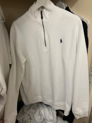 Ralph Lauren quartern-zip tröja - Vit långärmad tröja från Ralph Lauren med halv dragkedja och broderad logga på bröstet. Tröjan har hög krage och är tillverkad i ett mjukt, ribbat material som ger en clean och stilren look.