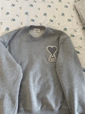 Grå sweatshirt från Ami Paris - Säljer en grå sweatshirt från Ami Paris med deras ikoniska hjärta-A-logga i tyg på bröstet. Tröjan har rund halsringning, ribbade muddar och är gjord i mjukt material som känns skönt mot huden. Perfekt för en avslappnad och trendig look.