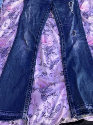 Blå bootcut jeans med slitningar - Säljer ett par blå bootcut jeans med slitningar och hål på framsidan. Jeansen har en klassisk femficksmodell och är tillverkade i denim med lätt tvättad look. Perfekta för dig som gillar en avslappnad och trendig stil.