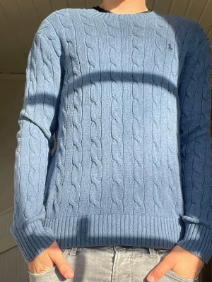 Blå kabelstickad tröja från Ralph Lauren - Snygg blå kabelstickad tröja från Ralph Lauren med rund halsringning och diskret logga broderad på bröstet. Tröjan har långa ärmar och ribbade muddar vid ärmslut och nederkant. Perfekt för dig som gillar klassisk och stilren look.