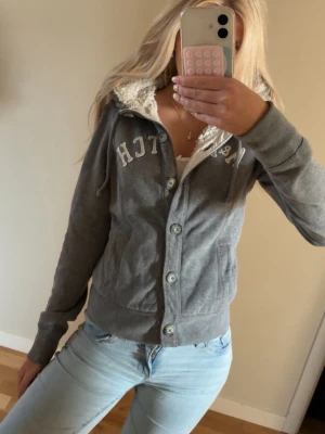 Grå hoodie med knappar och teddyfoder  - Mysig grå hoodie från Abercrombie & Fitch med knappar framtill och vitt teddyfoder i huvan. Hoodien har dragsko i huvan, fickor på sidorna och tryckt logga över bröstet. Perfekt för chill dagar och snygg till jeans. Storlek M men passar mig som har xs/s. Liten fläck finns men går bort i tvätten.