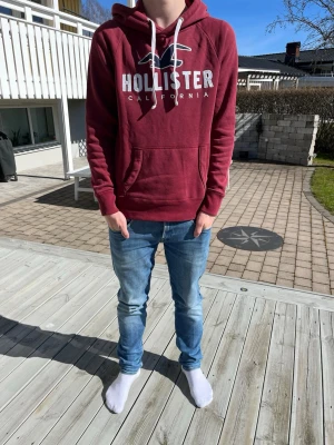 Vinröd hoodie från Hollister - Snygg vinröd hoodie från Hollister med stor vit logga och texten 'California' på bröstet. Klassisk känguruficka framtill och vita dragsnören i huvan. Perfekt för en avslappnad och trendig look.