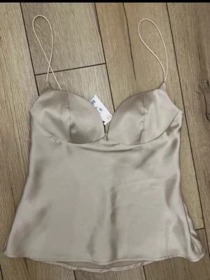 Champagnefärgad satin linnetopp - Snygg champagnefärgad linnetopp i satin med tunna axelband och lätt vadderade kupor. Toppen har en mjuk glansig finish och en feminin siluett som passar perfekt till både jeans och kjol. Enkel men elegant design som känns lyxig.