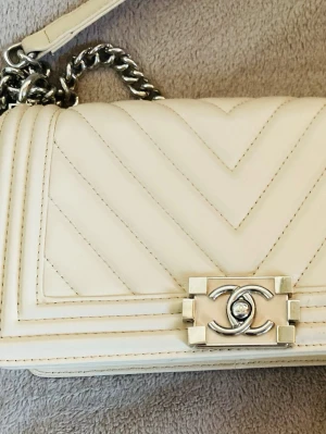 Vit Chanel Boy Bag i läder - Säljer en ikonisk Chanel Boy Bag i vitt läder med chevronmönster och silverfärgad kedjerem. Väskan har det klassiska CC-spännet framtill och en stilren, boxig form. Perfekt för dig som vill ha en lyxig och trendig accessoar.
