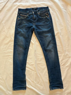 Blå slim fit jeans från Replay - Snygga blå slim fit jeans från Replay med klassisk femficksdesign och subtila slitningar. Jeansen har dragkedjegylf, silverfärgade knappar och detaljerade sömmar på bakfickorna. Perfekta för en trendig och avslappnad stil.