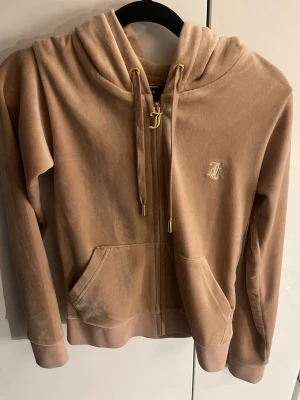 Beige velour hoodie från Juicy Couture - Säljer en beige velour hoodie från Juicy Couture med dragkedja, fickor framtill och guldfärgade detaljer på snörena. Ikoniskt JC-märke broderat på bröstet. Perfekt för en chill och trendig look.