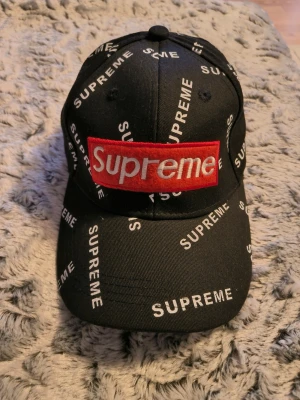Svart Supreme keps med logga - Säljer en svart keps från Supreme med vit logotyp tryckt över hela kepsen och en stor röd Supreme-patch framtill. Klassisk böjd skärm och justerbar rem bak. Perfekt för dig som gillar streetwear och vill sticka ut.