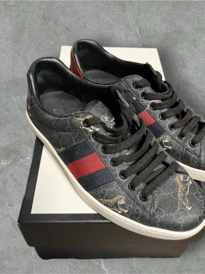 Gucci sneakers med tigerprint - Snygga Gucci sneakers i lågt modell med svart och grått monogrammönster, röda och marinblå ränder på sidan samt coola tigertryck. Skorna har svarta skosnören och vit sula. Materialet är canvas med läderdetaljer. Perfekta för dig som vill sticka ut med exklusiv streetstyle. Använda 1 gång nästan nya ej box