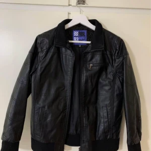 Svart skinnjacka från Rebel - Säljer en svart skinnjacka från Rebel med dragkedja och ribbade muddar. Jackan har en bröstficka med dragkedja och två sidofickor. Perfekt för dig som vill ha en klassisk och stilren look.