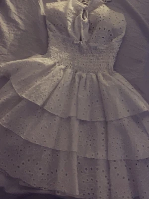 Vit volangklänning - Supersöt vit kortklänning med flera volanger och broderade spetsdetaljer. Klänningen har smock i midjan, knytning vid bysten och är ärmlös. Perfekt för sommarens alla tillfällen!