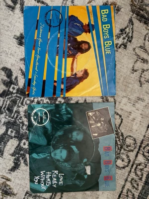 2 st Bad Boys Blue vinylsinglar - Två vinylsinglar med Bad Boys Blue: 'Heartbeat' i blått och gult omslag med bandfoto och 'Love Really Hurts Without You' i svart och turkost omslag med bandfoto och tecknad bild. Båda skivorna är i pappersfodral och har tydliga 80-talsvibbar. Perfekt för samlare av retro musik och vinyl.