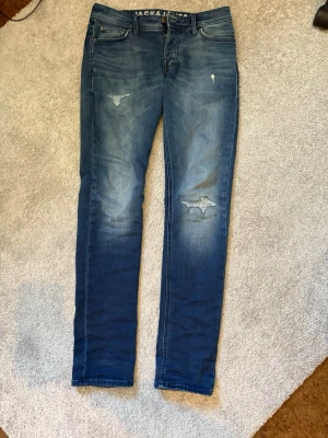 Blå slim fit jeans från Jack & Jones W30 L34 - Snygga blå slim fit jeans från Jack & Jones med slitningar och lagade detaljer på framsidan. Klassisk femficksmodell med normal midja och smala ben. Perfekta för en avslappnad och trendig look.