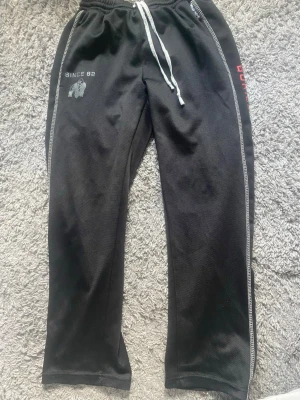 Svarta träningsbyxor Gorilla Wear - Säljer ett par svarta träningsbyxor från Gorilla Wear med vita kontrastsömmar och dragsko i midjan. Texten 'GORILLA WEAR SPORTSWEAR' i rött längs benet och logga framtill. Perfekta för gymmet eller sportaktiviteter.