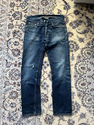 Blå slitna jeans från Our Legacy - Säljer ett par blå jeans från Our Legacy med snyggt slitna detaljer och hål på ena benet. Jeansen har klassisk femficksmodell, raka ben och är gjorda i denim. Perfekta för en avslappnad och trendig look.