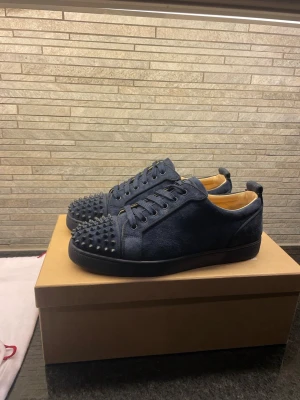  Äkta Mörkblå sneakers med nitar från Louboutin -  Äkta Snygga mörkblå sneakers från Christian Louboutin i mocka med matchande snörning och coola svarta nitar på tån. Skorna har en låg silhuett och svart sula. Perfekta för dig som vill sticka ut med en edgy och exklusiv stil.