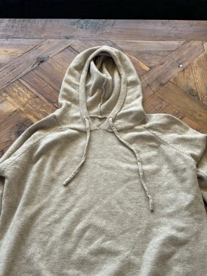 Beige hoodie med dragsko - Mysig beige hoodie med huva och dragsko. Tillverkad i mjukt stickat material och har ribbade muddar vid ärmslut och nederkant. Perfekt för en avslappnad och chill stil.
