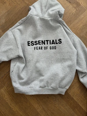 Grå Essentials Fear of God hoodie - Snygg grå hoodie från Essentials Fear of God med stor svart logga på ryggen. Klassisk passform med huva och ribbade muddar. Tillverkad i mjukt bomullsmaterial som känns skönt mot huden. Perfekt för en avslappnad streetwear-look.
