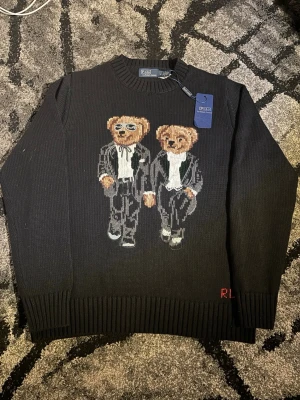 Svart stickad tröja Polo Bear Ralph Lauren - Svart stickad tröja från Polo Ralph Lauren med ikoniskt Polo Bear-motiv framtill där två björnar bär kostym. Rund halsringning och ribbade muddar vid ärmslut och nederkant. RL-broderi i rött nedtill. Perfekt för dig som gillar statement-plagg.