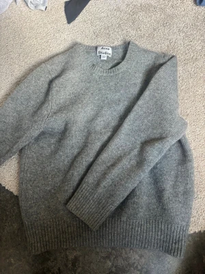 Grå stickad tröja från Acne Studios - Mysig grå stickad tröja från Acne Studios med rund halsringning. Bra skick förutom ett hål som jag sytt dit en annan tygbit på, se på bild 3. 