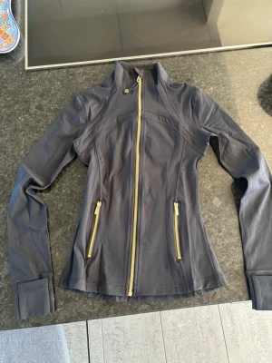 Svart vindjacka från Lululemon - Storlek 2 motsvarar xs/s. Snygg svart träningsjacka från Lululemon med figurnära passform och högt krage. Jackan har guldfärgade dragkedjor framtill och på fickorna samt tumhål vid ärmsluten. Perfekt för löpning eller gymmet. 