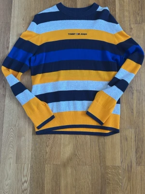 Randig stickad tröja Tommy Jeans - Säljer en stickad tröja från Tommy Jeans med breda ränder i gult, marinblått, ljusgrått och blått. Tröjan har rund halsringning och långa ärmar. Materialet är mjukt och stretchigt, perfekt för dig som gillar färg och streetstyle.
