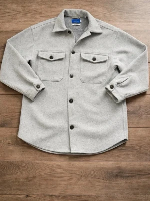 Ljusgrå overshirt från Weekday - Stilren ljusgrå overshirt från Weekday med två stora bröstfickor och svarta knappar. Overshirten har klassisk krage, raka ärmar med knapp vid ärmslut och är gjord i ett mjukt, tjockare material som ger en clean look.
