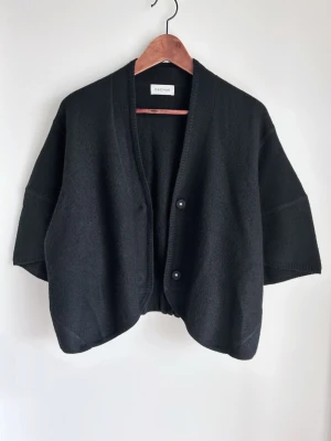 Svart kortärmad kofta från Dagmar - Stilren svart kofta från Dagmar med korta, vida ärmar och två knappar framtill. Koftan har en avslappnad passform och resår i midjan baktill för en snygg siluett. Tillverkad i mjukt material som känns lyxigt och bekvämt.