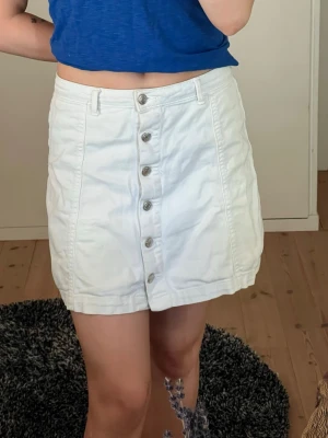 Vit kortkjol med knappar från gina tricot - Snygg vit kortkjol från Gina i bomull med raka linjer och knappar framtill. Kjolen har hällor för bälte och en enkel, clean look som passar till det mesta. Den har varit en favorit så använd mycket men inga större defekter på den. 