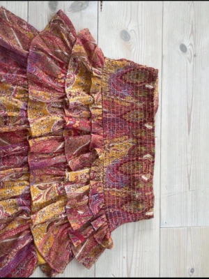 Mönstrad volangkjol i varma färger - Kort volangkjol med paisleyinspirerat mönster i rött, gult och lila. Kjolen har bred smock i midjan och flera lager volanger som ger en bohemisk vibe. Perfekt för dig som gillar färg och mönster!