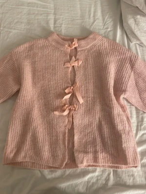 Rosa stickad tröja med rosetter - Supersöt ljusrosa stickad tröja med tre dekorativa rosetter framtill. Tröjan har en mjuk, fluffig struktur och ribbad halsringning. Perfekt för dig som gillar gulliga detaljer och en mysig känsla. Materialet känns som en mjuk ull- eller mohairblandning.