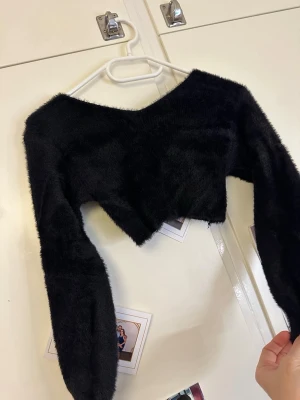 Svart fluffig croppad topp - Säljer en svart, supermjuk och fluffig croppad topp med lång ärm. Toppen har en bred, asymmetrisk halsringning och är perfekt för dig som vill ha en statement piece i garderoben. Materialet känns som syntetpäls och ger en lyxig känsla.