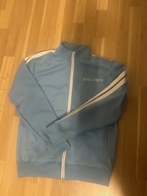 Ljusblå zip hoodie från Palm Angels - Säljer en ljusblå hoodie från Palm Angels med vit dragkedja och vita ränder längs ärmarna. Klassisk streetwear-look med hög krage och logga på bröstet. Perfekt för dig som gillar stilrena och sportiga plagg.