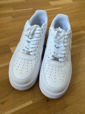 Nike Air Force 1 vita sneakers - Klassiska Nike Air Force 1 sneakers i helvitt läder med perforerad tåbox och vit sula. Swoosh-logga på sidan och vita skosnören. Tidlös och clean design som passar till det mesta. Perfekt för dig som gillar streetwear och vill ha en ikonisk sneaker i garderoben.