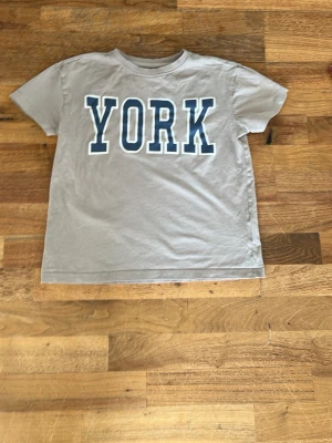 Beige t-shirt med tryck 'YORK' - Snygg beige t-shirt med stort blått tryck 'YORK' på bröstet. Klassisk rund halsringning och korta ärmar. Perfekt till jeans eller shorts för en avslappnad look.
