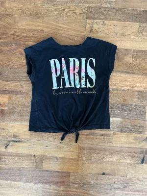 Svart knyt-topp med PARIS-tryck - Svart topp med stort PARIS-tryck i blommigt mönster framtill och texten 'l'amour is all we need' under. Toppen har korta ärmar och knytning nedtill för en avslappnad look. Perfekt för dig som gillar trendiga statement-plagg.