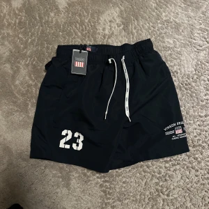 Svarta badbyxor från Vanson Drive - Svarta badbyxor med vit snörning i midjan, tryckt siffra 23 på vänster ben och logga med text på höger ben. Klassisk modell med elastisk midja och sportig look. Materialet är troligtvis snabbtorkande syntet.