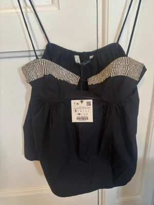 Svart linnetopp med strass från Zara - Trendig svart linnetopp från Zara med tunna axelband och glittriga strassdetaljer längs överdelen. Toppen har en lätt puffig form och en liten cut-out framtill. Perfekt för dig som vill sticka ut med en snygg och glamorös look.