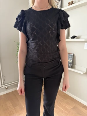 Svart blus med volangärmar - Snygg svart blus med diskret rutmönster och volangdetaljer på ärmarna. Blusen har rund halsringning och är tillverkad i ett lätt, något glansigt material som ger en elegant känsla. Perfekt att styla med både jeans och kostymbyxor.