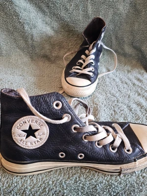 Svarta Converse All Star Highs - Klassiska svarta Converse All Star high-top sneakers i läder med vita gummisulor och tåhättor. Skorna har vita snören och metallöljetter samt den ikoniska Converse-loggan på sidan. Perfekta för en cool och avslappnad stil.