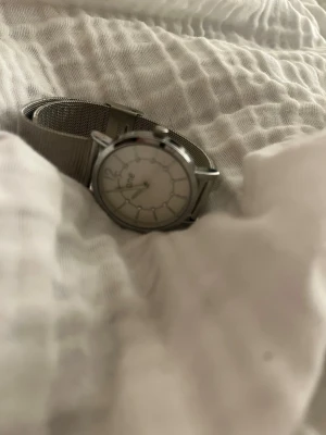 Silverfärgad klocka från Guldfynd - Stilren klocka från Guldfynd med silverfärgat mesharmband och rund vit urtavla. Klockan har minimalistisk design med tydliga markeringar och metallboett. Perfekt accessoar för dig som gillar klassisk och enkel stil.