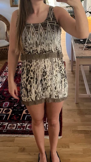 Kortklänning  - Kortklänning med coolt mixat mönster i beige, svart och brunt. Överdelen har zigzag-mönster och kjolen har leopardliknande print. Midjan och nederkanten är dekorerade med bred spets. Ärmlös och luftig modell, perfekt för varma dagar.
