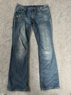 Bootcut jeans  - Säljer ett par blå jeans från Vigoss köpta i London. Dem är i Bootcut modell och har slitningar framtill. Baksidan har snygga fickor dekorerade med nitar och knappar. 