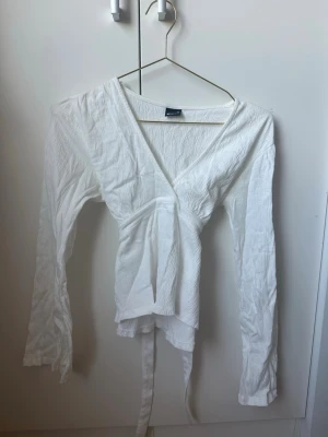 Gina vit omlottblus med knytband - Snygg vit omlottblus med långa ärmar och knytband i midjan. Blusen har en djup v-ringning. Perfekt att styla med jeans eller kjol för en trendig outfit. Från Gina storlek XS. Använd typ 2 ggr så i perfekt skick.