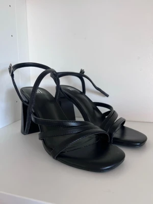 Svarta klackar med blockklack XIT - Stilrena svarta sandaletter från XIT med bred blockklack och fyrkantig tå. Skorna har tunna remmar i konstläder som korsar över foten och justerbar ankelrem med spänne. Aldrig använda endast testade hemma. 