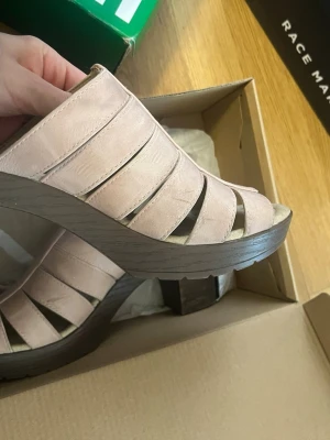 Ljusa platåsandaler med klack - Säljer ett par ljusrosa sandaler med breda remmar och öppen tå. Skorna har en hög platåklack i trälook och är tillverkade i ett material som liknar mocka. Perfekta för dig som vill ha både höjd och stil i sommar.