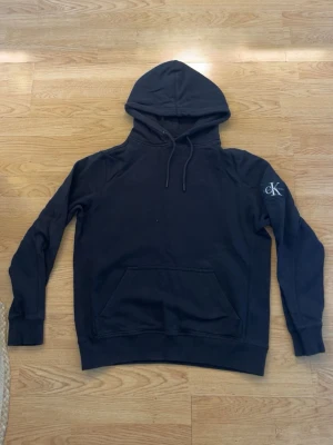 Calvin Klein hoodie - Snygg svart hoodie från Calvin Klein med klassisk huva och dragsnören. Stor magficka framtill och CK-logga broderad på vänster ärm. Tillverkad i mjukt bomullsmaterial som känns skönt mot huden. Perfekt för en avslappnad och stilren look. Inga hål eller fläckar.