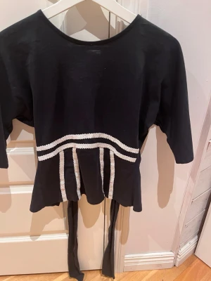 Svart topp med knyt och vita detaljer - Cool svart topp med halvlånga ärmar och vita dekorativa band framtill. Toppen har knytband i midjan och öppen rygg för en trendig look. Perfekt för dig som gillar unika detaljer och vill sticka ut.